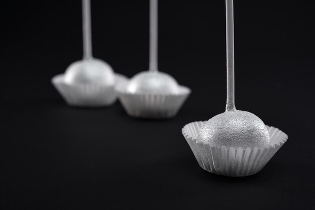 Closeup of three glazed sweet silver cake pops on long sticks in white paper baskets. Delicious fresh candies isolated on black background. Concept of desserts, confectionary.