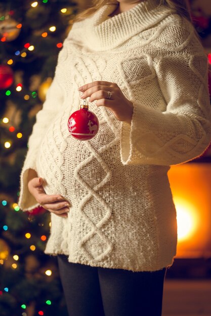 Closeup toned photo of pregnant woman in wool sweater holding red Christmas decorative ball