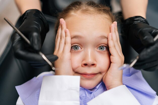 Closeup top view portret van schattig bang kind meisje dat hoofd houdt van angst met hand zit op tandarts stoel Closeup handen van onherkenbare tandarts die roestvrijstalen tandheelkundige gereedschappen bij gezicht houdt