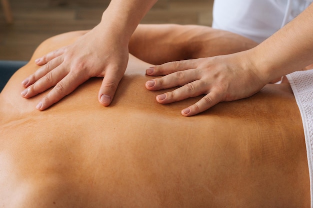 Primo piano vista dall'alto di un massaggiatore maschio irriconoscibile con forti mani tatuate che massaggiano la schiena di uno sportivo muscolare irriconoscibile sdraiato sullo stomaco