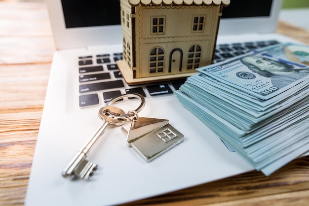 Крупный план ключей от игрушечного деревянного дома и долларов на ноутбуке Концепция покупки ипотечного кредита на аренду дома