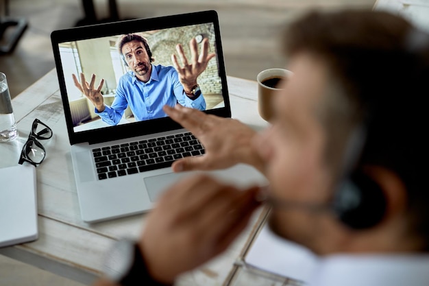 Photo closeup of uncertain businessman gesturing while having video call with a colleague over laptop focus is on computer screen