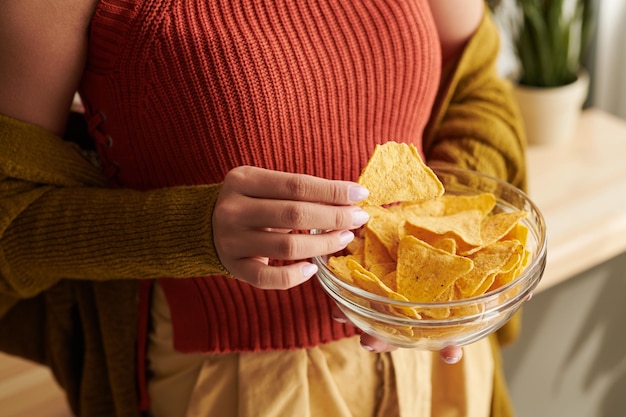 Closeup of unrecognizable woman in bright top and cardigan eating nachos from bowl at home unhealthy food concept