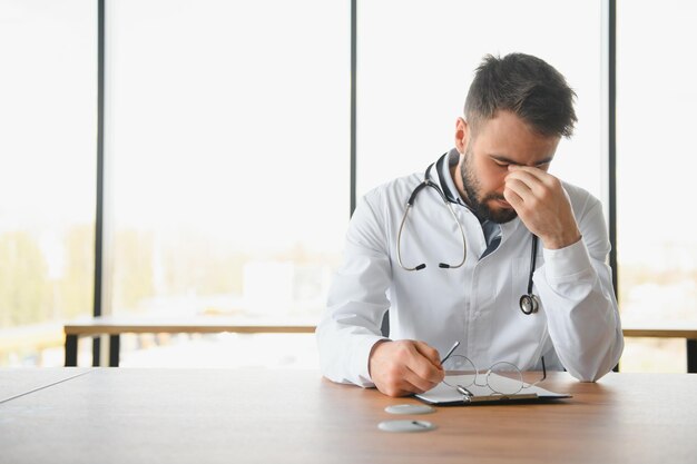 Photo closeup of upset tired medical worker closing head with hands depressed male doctor sitting in office stethoscope on persons neck modern medicine and healthcare concept