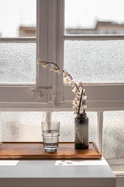 Photo closeup of a vase with flowers and a glass stands on a wooden tray on a window in a hotel room