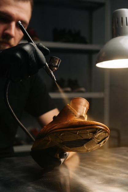 Closeup vertical shot of bearded shoemaker wearing black gloves spraying paint of light brown leather shoes closeup