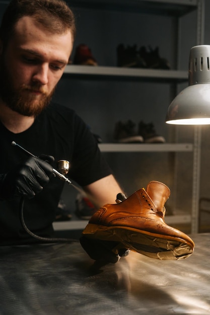 Closeup vertical shot of bearded shoemaker wearing black gloves spraying paint of light brown leather shoes closeup