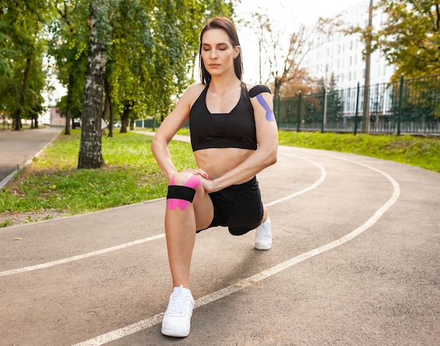 Closeup view of bruette flexible woman with muscular body warming up outdoors, practicing deep lunges. Stunning fit young sportswoman training in summer park, wearing kinesiology tapes on knees.
