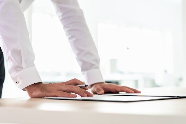 Closeup view of businessman leaning on his office desk holding a pen to sign a contract or document in a folder.