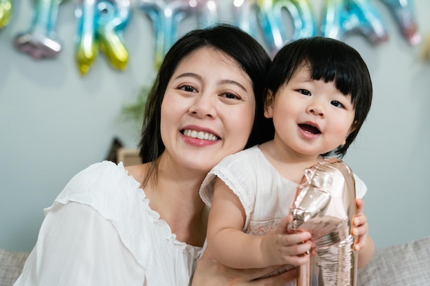 생일 파티에서 웃는 얼굴로 카메라를 바라보면서 사랑스러운 아기 딸을 안고 있는 쾌활한 아시아 엄마의 클로즈업 보기