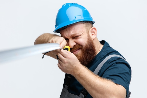 Closeup view of concentrated young male construction worker wearing safety helmet and uniform stretching measuring tape towards camera with one eye closed isolated on white background