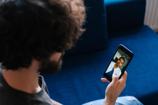 Closeup view from back to sick young male patient chatting via\
video call with africanamerican doctor on mobile phone from\
home