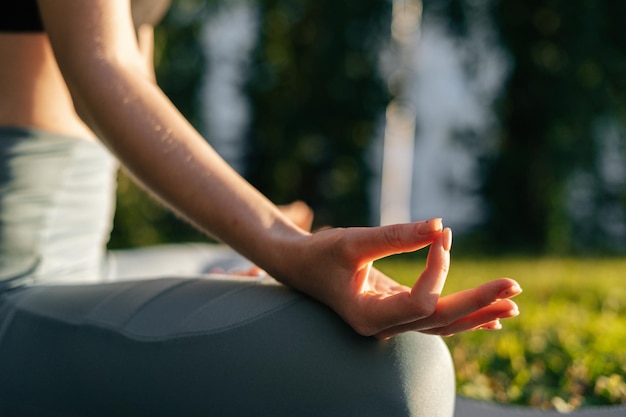 Closeup view of hands of young woman holding fingers on knee in yoga mudra position while sitting at lotus pose on green grass Unrecognizable female enjoying meditation on park in morning at dawn