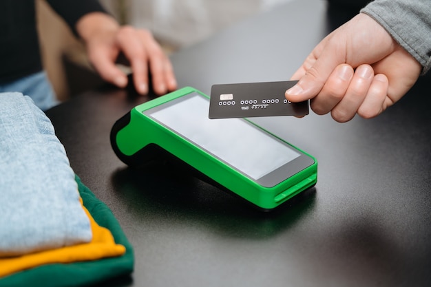 Vista del primo piano, cliente maschio che utilizza la carta di credito bancaria per il pagamento contactless dal terminale nfc sul bancone mentre si fa la spesa nel negozio di abbigliamento