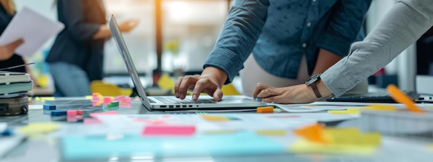 closeup view of a persons hands working on a laptop with other individuals in the background surrounded by colorful sticky notes papers and a collaborative work environment