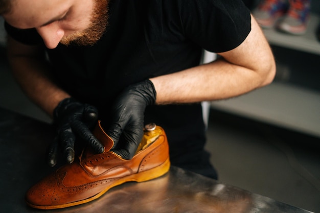 Photo closeup view of shoemaker in black gloves rubbing paint on light brown leather shoes with fingers