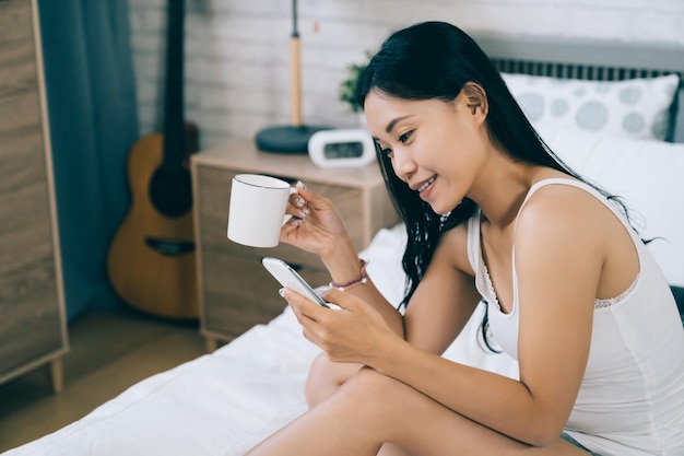 closeup view of smiling asian young lady browsing friends’ stories on social networking website while having morning coffee. technology and lifestyle.