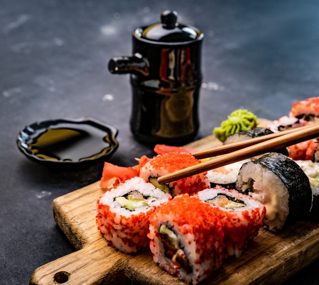 Closeup view of sushi maki set served on wooden tablet with soy sauce in bowl and person hand holdin...