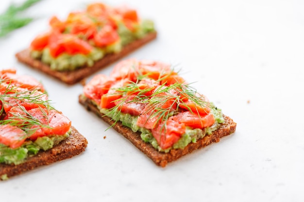 Closeup view of three sandwiches with rye bread avocado and smoked salmon on a white kitchen table Copy space