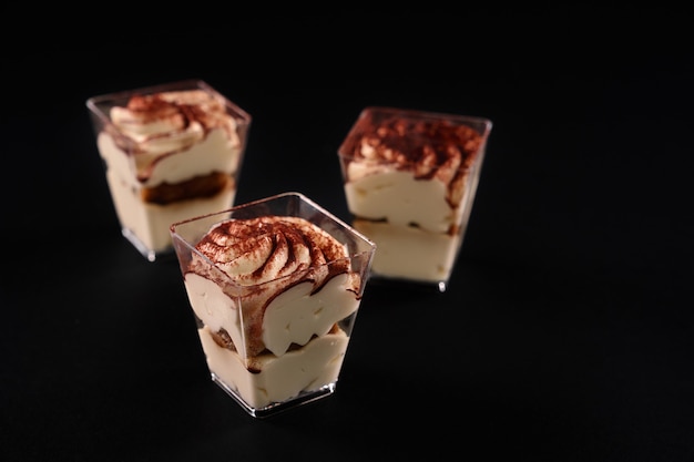 Vista ingrandita di tre piccoli vetri con dessert in fila decorata con panna montata e cacao, isolato sul nero. torta tiramisù con tre strati di biscotto al cioccolato e mousse di sciroppo naturale al caffè.