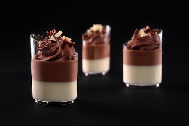 Closeup view of three small glasses with creamy layered chocolate and milk panna cotta. Sweet delicious dessert decorated with brown whipped cream topping, isolated on black background. Food concept.