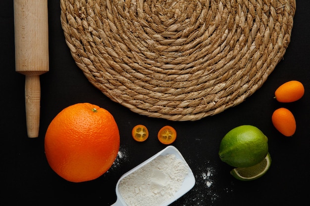 Closeup view of trivet with lime orange tangerine kumquat flour in measure spoon with rolling pin on black background