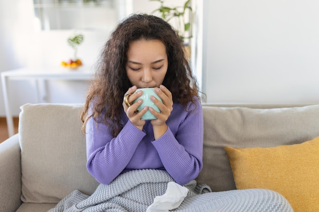 Closeup view of young Asian woman with cup of hot drink at home covered with blanket People drinks and leisure concept young woman with cup of tea or coffee at home
