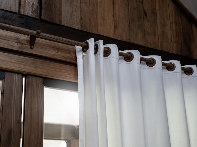 熱帯の切妻の建物の木の板の壁に飾られた木製のカーテン レール バーの白い壁カーテンのクローズ アップは、外からの日光で部屋の中のガラスの引き戸の近く