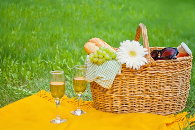 Closeup of wine glasses on the yellow cover, picnic basket with food and flower on the green grass