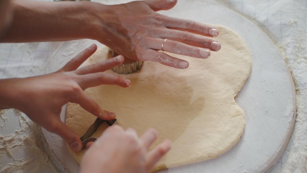 Closeup woman and girl hands cutting dough with cookie cutters on kitchen table Top view of mother and daughter cooking biscuits together on domestic kitchen in slow motion