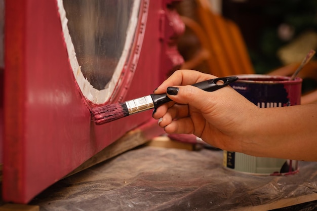 Closeup of woman hand with brush painting carved ornaments on lower part of door of antique cupboard made of wood in pink color Giving new life to old stuff Hand restoration of old furniture