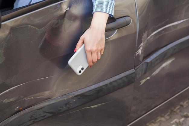 Photo closeup of woman hand with mobile phone hanging from automobile window, drunk driver in damaged car, injured female in automobile with dents and scratches, help for aggrieved. outdoor shot.
