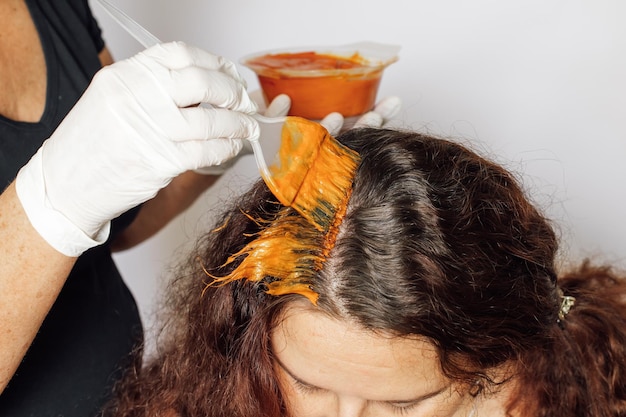 Foto primo piano delle mani della donna in guanti bianchi con la colorazione della spazzola radici dei capelli grigi della donna su sfondo bianco colorazione dei capelli a casa sbarazzarsi dei capelli grigi antiage