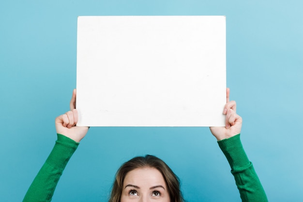 Closeup woman holding a white board copy space above her head against blue wall