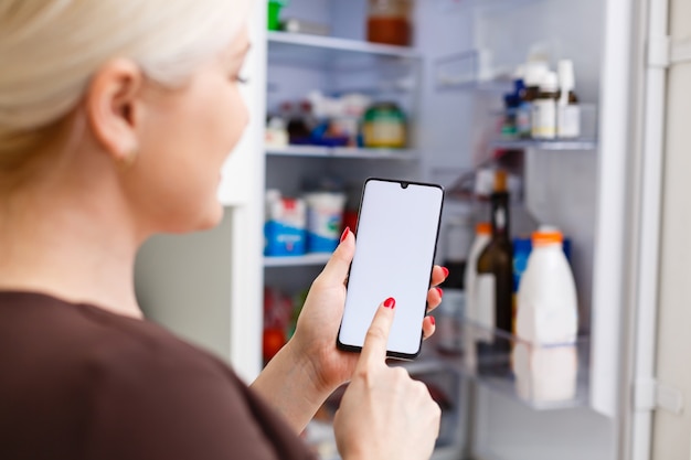 自宅の冷蔵庫に対して携帯電話のエネルギーラベルを使用して女性の手のクローズアップ