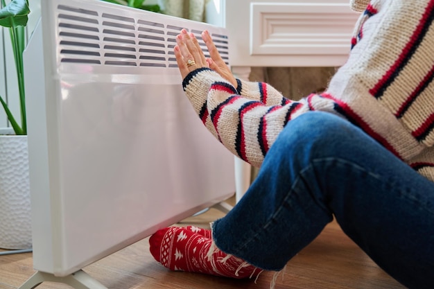 closeup of a woman's hands legs warming near an electric heating radiator heating cold season autumn winter comfort energy saving crisis home lifestyle concept