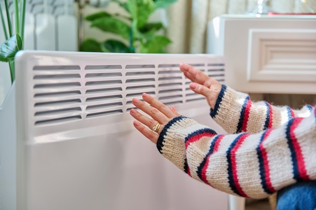 Closeup of a woman's hands warming near an electric heating radiator Heating cold season autumn winter comfort energy saving crisis home lifestyle concept