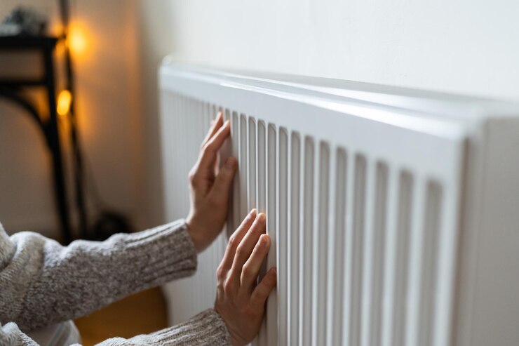 Photo closeup of woman in sweater puts hands on room central heating battery to warm up in cold apartment