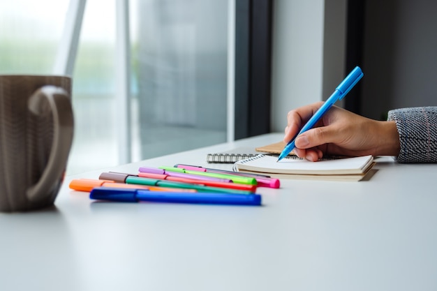 Closeup  of a woman writing on a blank notebook with colored pens and coffee cup on the table