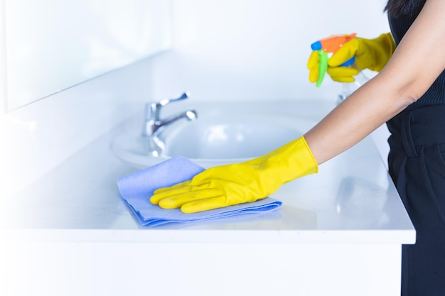 Closeup women hand maid or charwoman cleaning modern new basin in bathroom Water tap clean using yellow gloves and blue cloth with foggy sanitizer spray