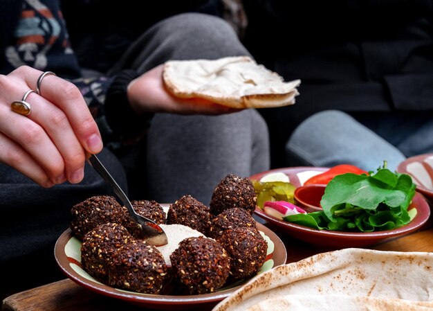 Foto close-up di mani di donne che tengono il pane e spargono l'hummus un piatto di falafel e verdure