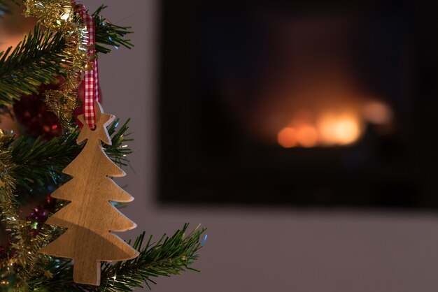 Closeup of wooden holiday ornament hanging on Christmas tree with fire in fireplace in background.