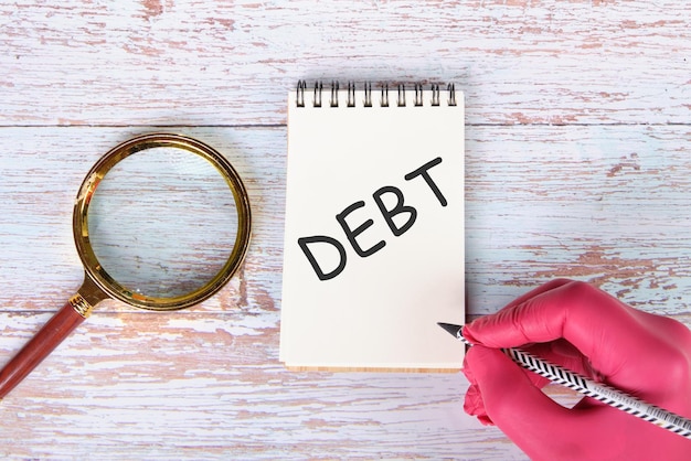 Closeup of the word DEBT on a notebook written by a hand in a red glove