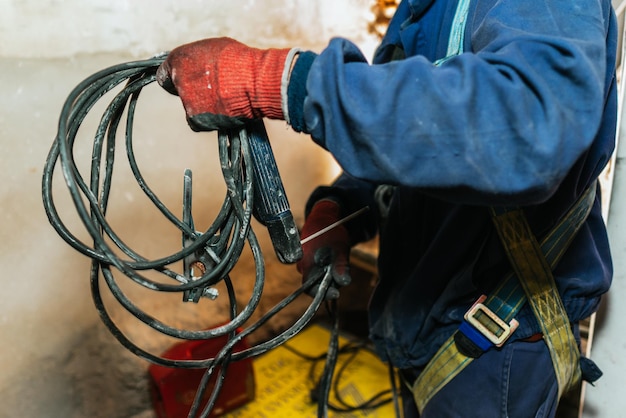 Photo closeup of a worker in a blue uniform, red gloves and a harness holding a dirty black cable