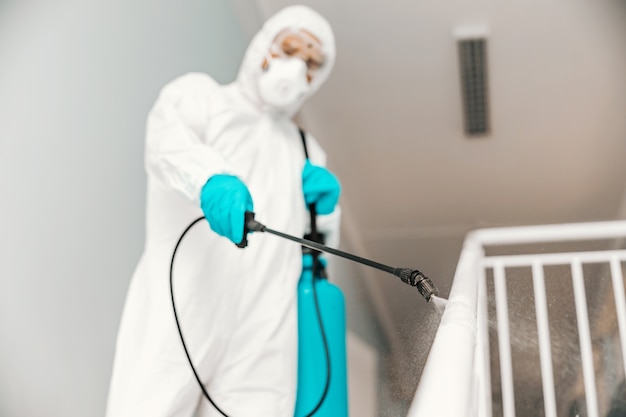 Closeup of worker in sterile uniform, with gloves and facial mask sterilizing railing in school