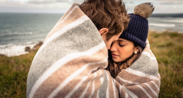 Photo closeup of young beautiful couple embracing under blanket in a cold day with sea and dark cloudy sky on the background