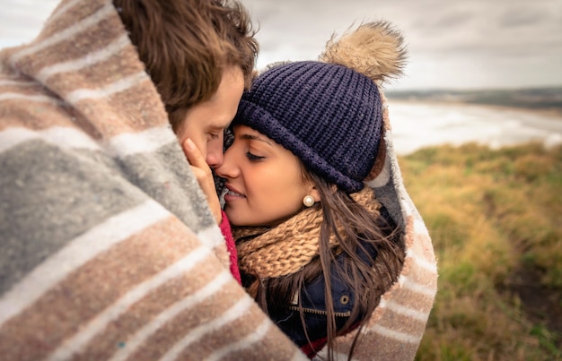 Closeup of young beautiful couple embracing under blanket in a cold day with sea and dark cloudy sky on the background