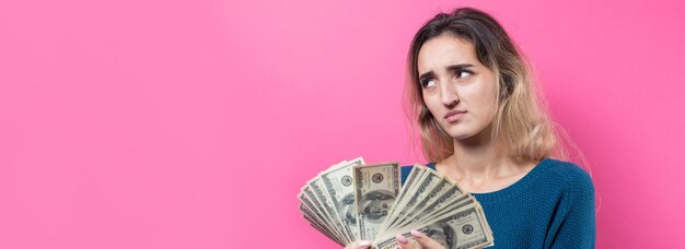 ピンクの背景の上にアメリカドルのお金を手にメガネの青いセーターで若い美しい女性のクローズアップ感情と喜びのジェスチャーを表現します