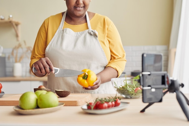 Closeup of young black woman cooking healthy meal in kitchen and recording video with smartphone cop