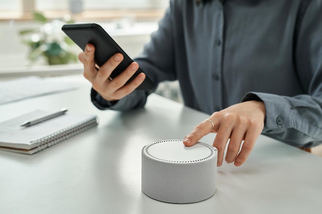 Closeup of young businesswoman connecting smart speaker with mobile phone sitting at desk